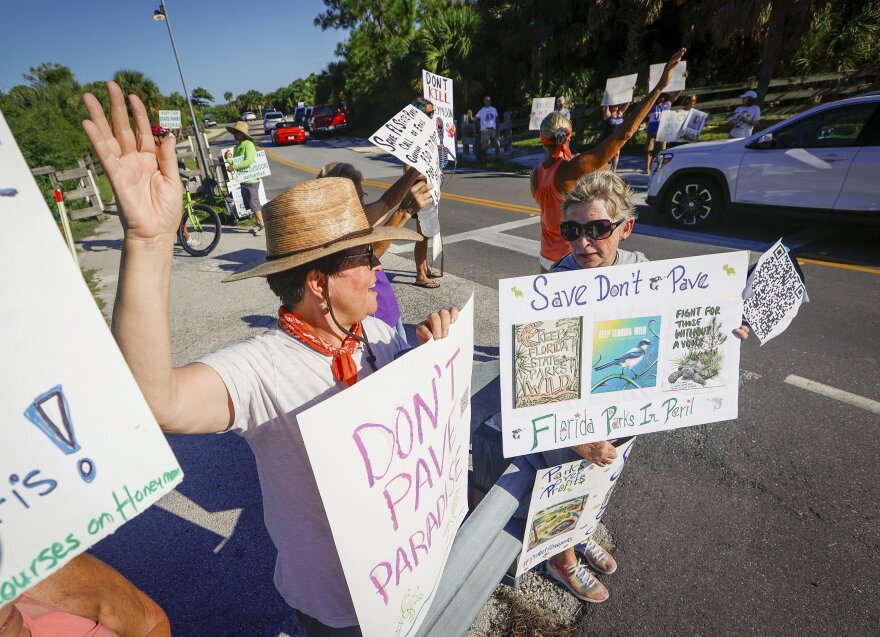 Michelle Birnbaum, 59, left, and Kathie Nutter, 67, right, both of Dunedin, hold signs and wave to drivers at the entrance to Honeymoon Island State Park during a rally Sunday, Aug. 25, 2024 in Dunedin, Fla. (Chris Urso/Tampa Bay Times via AP)
