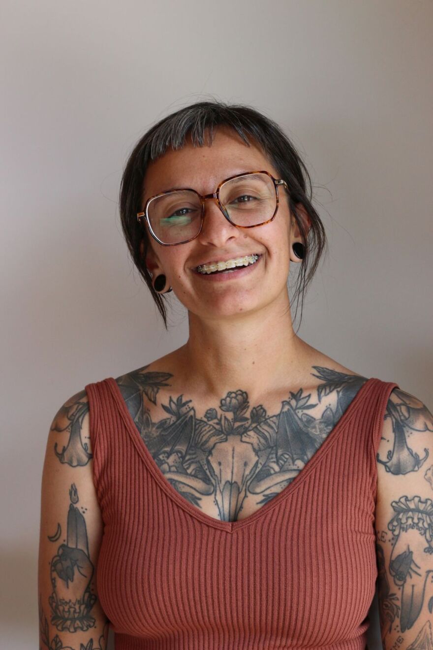 Dima Khalifa smiles. She has floral ttatoos across her chest, grazing her collarbone, and shoulders and wears an orange ribbed v-neck tank top.  