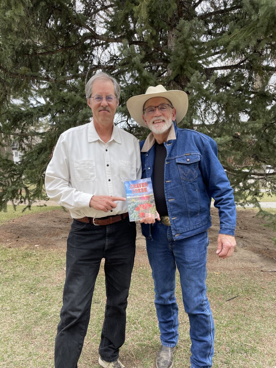 Two older men stand side-by-side, smiling at the camera. One is holding a book and pointing at it.