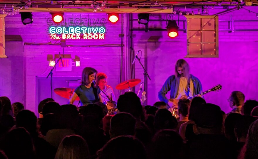 A large crowd of people stand in front of a stage at a music venue watching three band members perform.