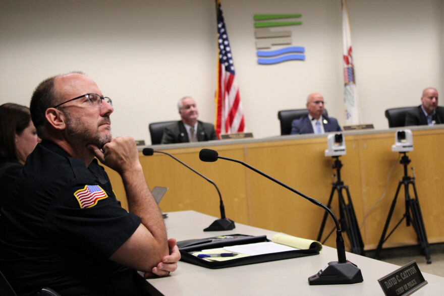 A police officer in uniform sits at a table during a meeting, with three other officials seated behind him in front of American and organizational flags.