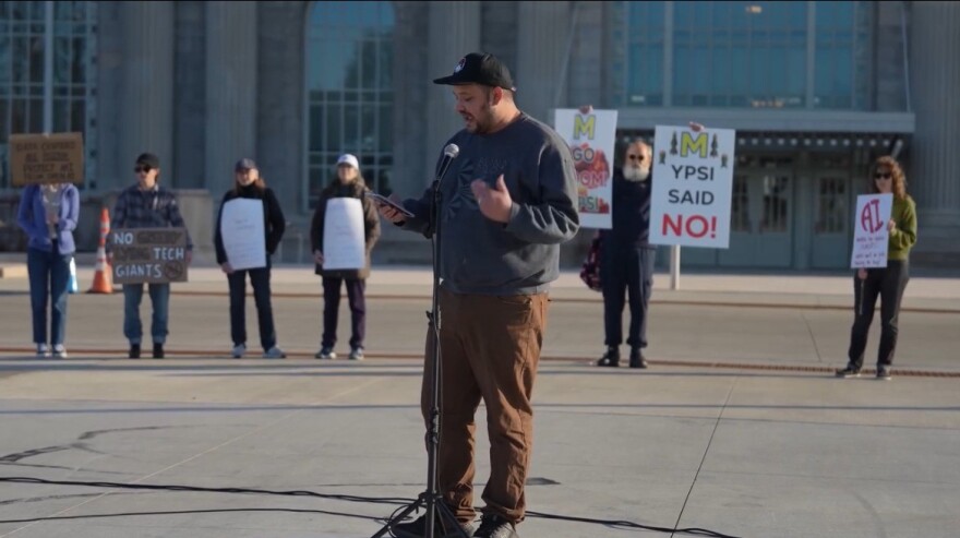 Michigan State Rep. Dylan Wegela speaks into a microphone. People stand behind him holding signs.