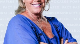 A professional headshot of a smiling woman with blonde hair and red-framed glasses, wearing a bright blue top with arms crossed, against a light blue-grey background with faint text.