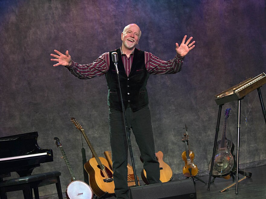 Man standing on stage with hands raised in front of a row of guitars