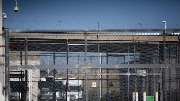 A chain-linked fence with barbed wire across the top with trucks parked off in the distance.