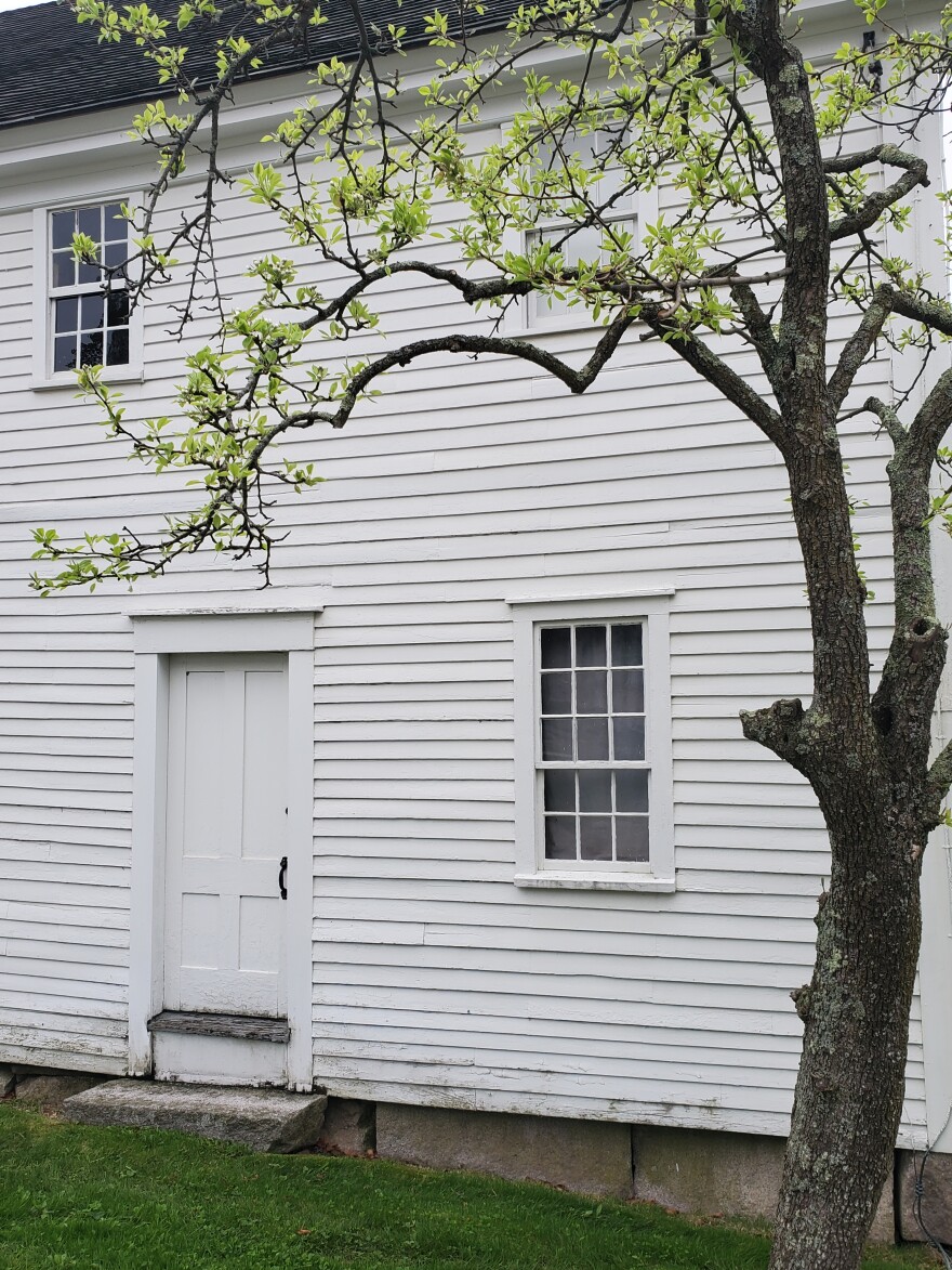 A white-slatted Shaker building next to a tree.