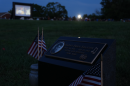 A grave marker with Harry Dean Stanton's portrait and name engraved, flanked by American flags. One of his films plays on a large screen nearby.