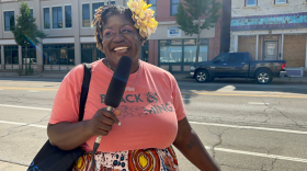 A Black woman standing on the sidewalk, holding a microphone and laughing while getting her picture taken. 