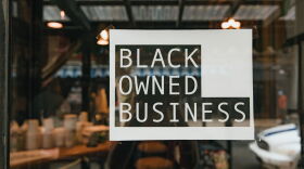 Black owned business sign taped on a window, seen from outside a store.