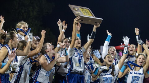 Quarterback Emme Albright is lifting up the plaque declaring them state champions alongside her St. James team. They are all in their blue and white uniforms and many of them have medals around their necks and in their hands showing off the win. They are jumping in celebration.
