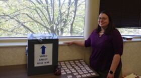 A woman in a purple long sleeve blouse and black pants stands to the right of a brown table. On top of the table sits the grey ballot deposit box with a sign signifying that it is indeed where a voter puts there ballot. A small cup of glue sticks sits in front of the box, with numerous "I Voted" stickers sitting on the table to its right. 