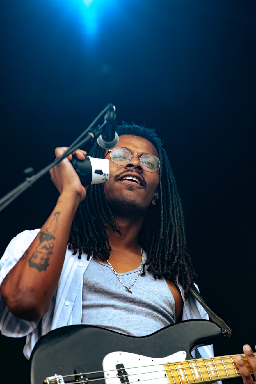 Caleb Buchanan, the lead guitar player for the indie act spilltab, stands on the Hinterland music festival mainstage with a black and white fender guitar around his neck. He is a Black man with locks wearing a white tank top under a stripped, short sleeved, button down blue shirt. 