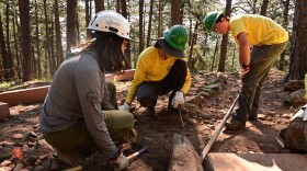 Three Boulder Junior Ranger Program participants wear hard hats, safety glasses and work gloves. They're using a variety of tools to clean up a rock-lined area of a park.