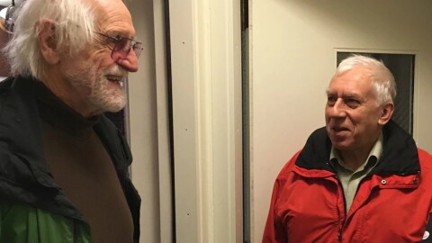Man in a green coat and brown sweater standing in a doorway speaking to a man in a red coat in front of an interior door.