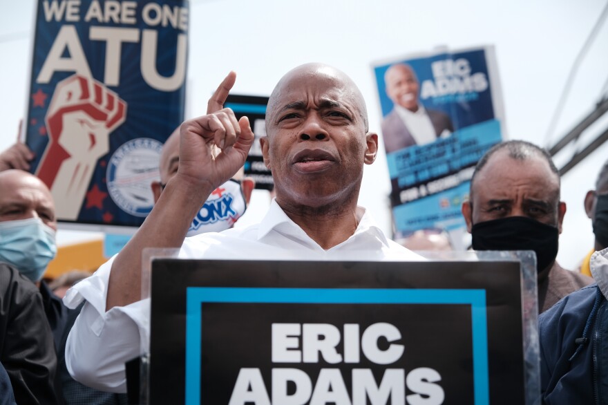 Eric Adams, wearing a white collared shirt, points a finger upward while speaking in a crowd behind a sign with his name on it.