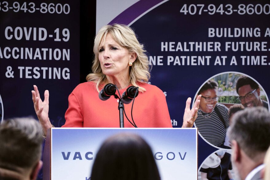 Jill Biden wears a coral-colored suit as she speaks into a microphone at an event.