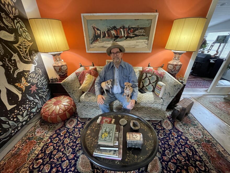 A man sits on a couch in between two matching lamps. Two small dogs sit on his lap.