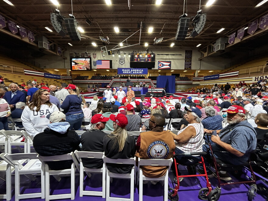 A large crowd of people seated in a high school gymnasium with a large sign in the back that reads "Make America Great Again."