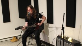 A seated musician with long hair and red glasses playing guitar and singing into a microphone in a radio studio. A small suitcase and an amp are to their right.
