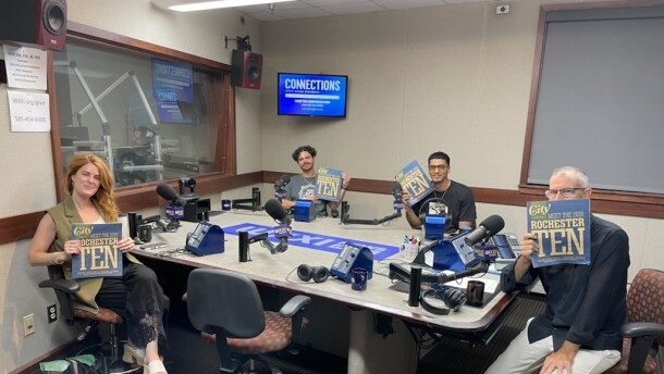 Four smiling people holding copies of CITY Magazine as they sit around a table in a radio talk studio: a woman front left has long wavy red hair and is wearing a green top, black pants, and white sandals; a man back left has short curly brown hair and is wearing a gold chair and a blue graphic t-shirt; a man back center has short dark hair and is wearing glasses, a gold chain, and a black graphic t-shirt; a man at right has very short dark hair and is wearing a black button-down shirt, tan linen pants, and grey and blue sneakers