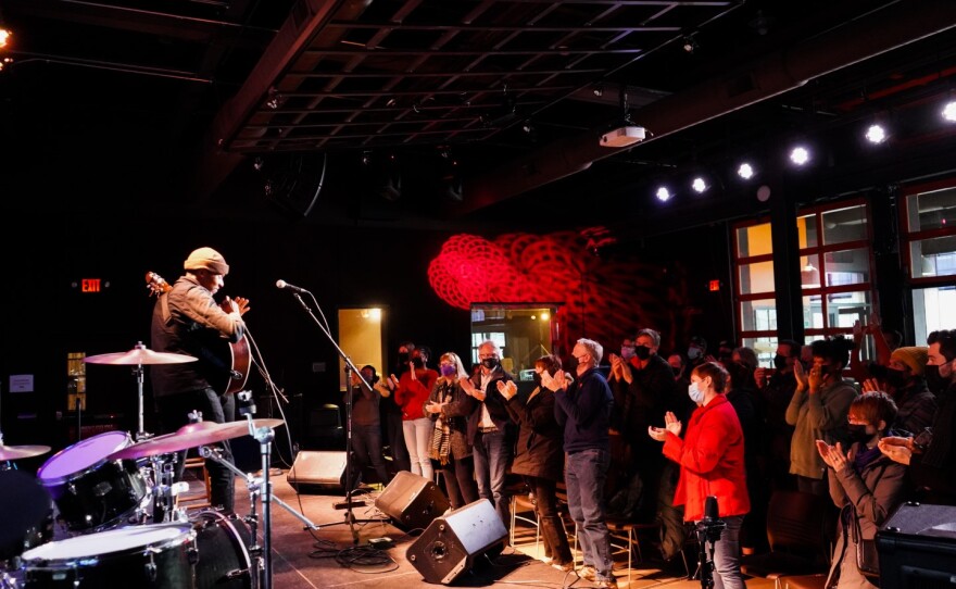 An audience in a crowded room stands and applauds as a man holding an acoustic guitar on stage acknowledges them.