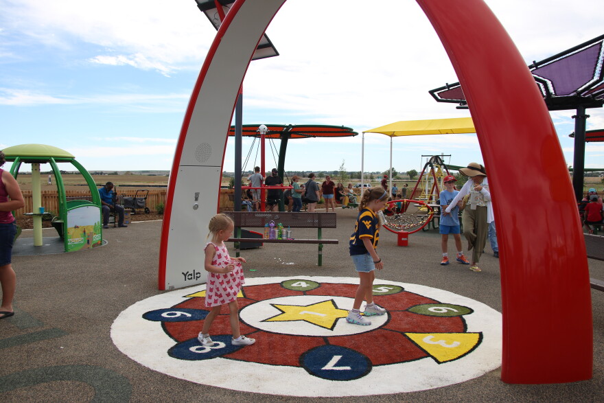 Two young girls -- one in a pink and white dress, and the other in a blue and yellow jersey -- hop and walk across colorful numbers on the ground. They are standing underneath a large red and white arch. Behind them is a playground.