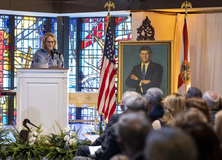 A woman speaks at a podium near a portrait of a man. 