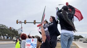 Students stand on the barrier at the edge of an intersection carrying signs that say "Love Melts ICE. Support our Immigrants" and "Skipping Lessons to Teach One."