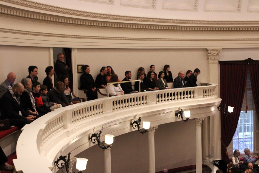 About 30 people sit on a balcony behind a white bannister.