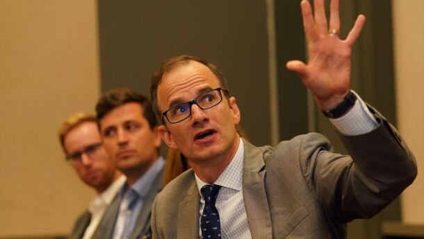 A man in a suit gestures at something while speaking, with multiple other people in suits sitting behind him.