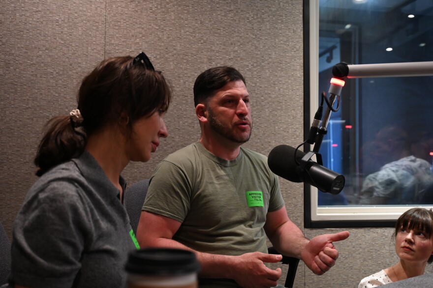 A man speaks into a microphone inside a studio. In the foreground is a woman with a pony tail 