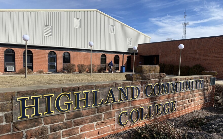 A short brick wall holds a sign that reads "Highland Community College" on it. Behind it is a brick building with a metal roof. A person is walking toward it on a sidewalk.