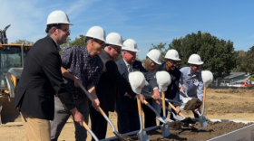 Seven people wearing white construction hats and holding shovels stand in front of a raised box of soil