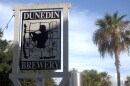 A black-and-white sign stands against a clear blue sky and a line of palm trees. A grey brewery canister is also visible in the background. The sign reads DUNEDIN BREWERY, and an engraved image shows the silhouette of a woman playing bag pipes. 