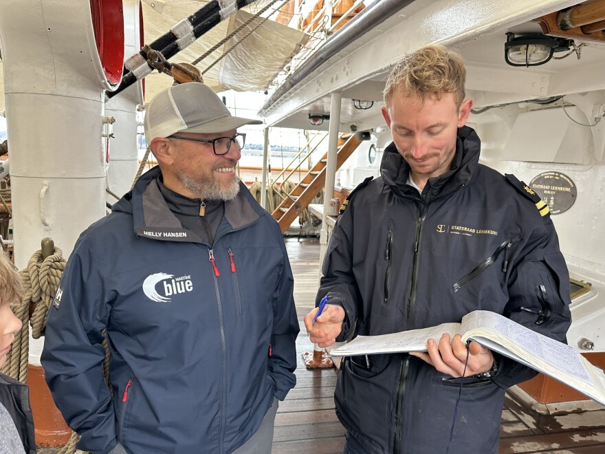 Two people on board a tall ship. One is holding a ledger and looking at it, and the other is turning to him and smiling.