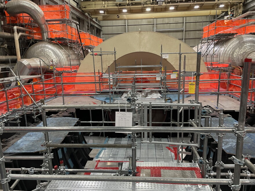 Metal scaffolding and bright orange netting cover the room, while the light-brown metal arch of the partially dissassembled turbine cover sits inside the Palisades nuclear plant.