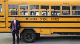 A man stands in front of a yellow Anchorage School District bus wearing a suit with a red tie.
