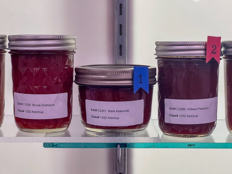 Three jars of homemade ketchup sit on a well-lit glass shelf. 