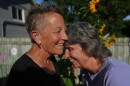2 older white women laugh in the backyard of a home  , the one on the left has short cropped grayish hair in a black top.... the one on the right has darker hair and a light blue top 