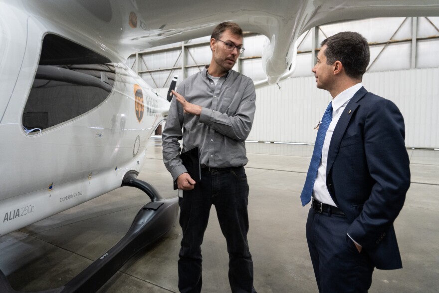 A man in a grey shirt and black pants stands next to a white aircraft, speaking with a man in a navy suit and tie.