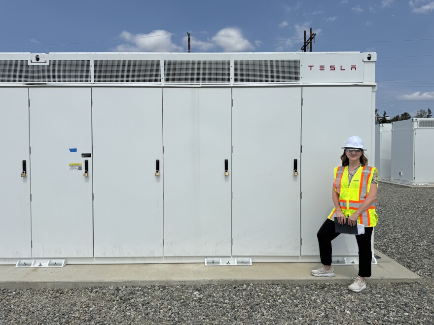 A woman in a bright yellow safety vest and white hard hat stands smiling alongside a row of vented white lockers that contain energy storage batteries.