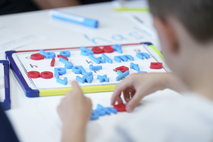 A young child, seen from behind sitting at a school table with a puzzle full of red and blue letters, tries to make words.