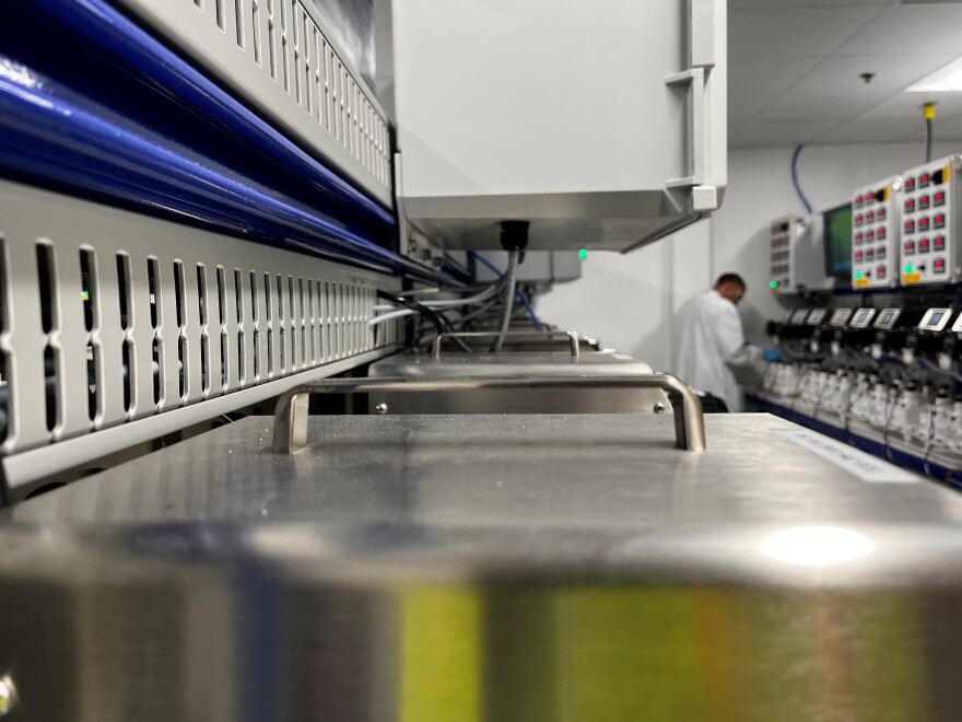 A metal machine in the foreground with a man in a white lab coat studying another machine in the background