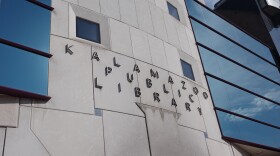 Silver letters on the side of a building read "Kalamazoo Public Library." The glass windows reflect the open blue sky on a bright sunny day.