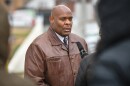 A man wearing a brown leather jacket talks in front of a microphone outdoors. There are out of focus people standing in the foreground, framing him.