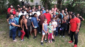 Group shot of multiple people standing on grass with trees behind them. 