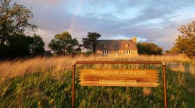 Beautiful photo of prairie with an old stone cabin. There is a faint rainbow overhead. In the foreground there's a sign that says Iowa Lakeside Laboratory established 1909.