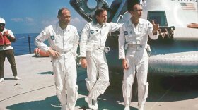 Three men in white jumpsuits stand in front of a spacecraft. They appear to be talking with someone out of frame to their left.