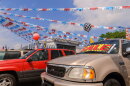 A vibrant car dealership scene with several vehicles parked in front. Colorful banners and flags, including red, silver, and blue streamers, are strung overhead. A prominent "SALE" sign is displayed on the windshield of a beige SUV. In the background, there is a small building adorned with a racing-themed checkered pattern.