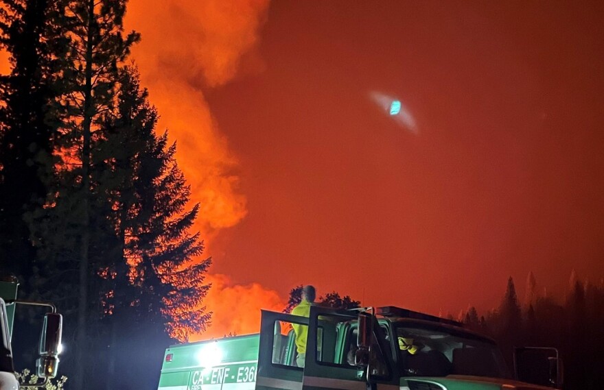 The sky is red and there's smoke coming from behind a silhouetted tree. There is a firefighting person standing in a truck facing the smoke.